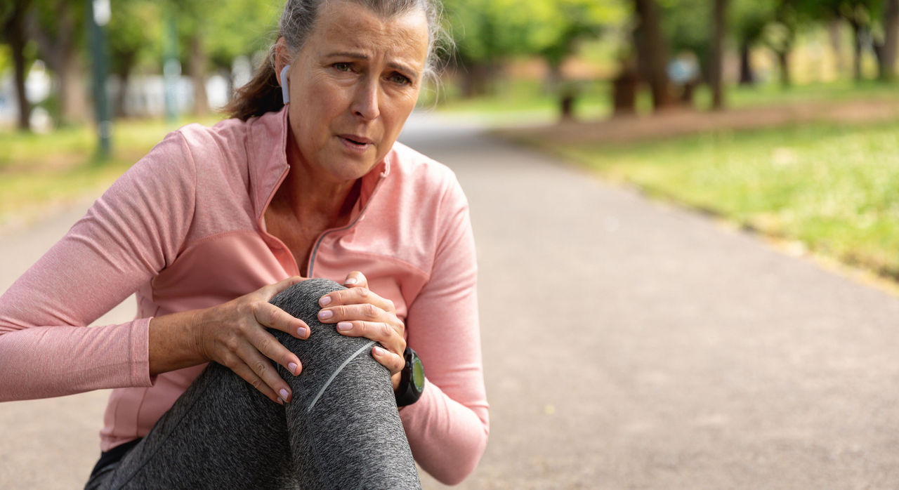 A middle-aged jogger sits on a running path, clutching her knee in pain.