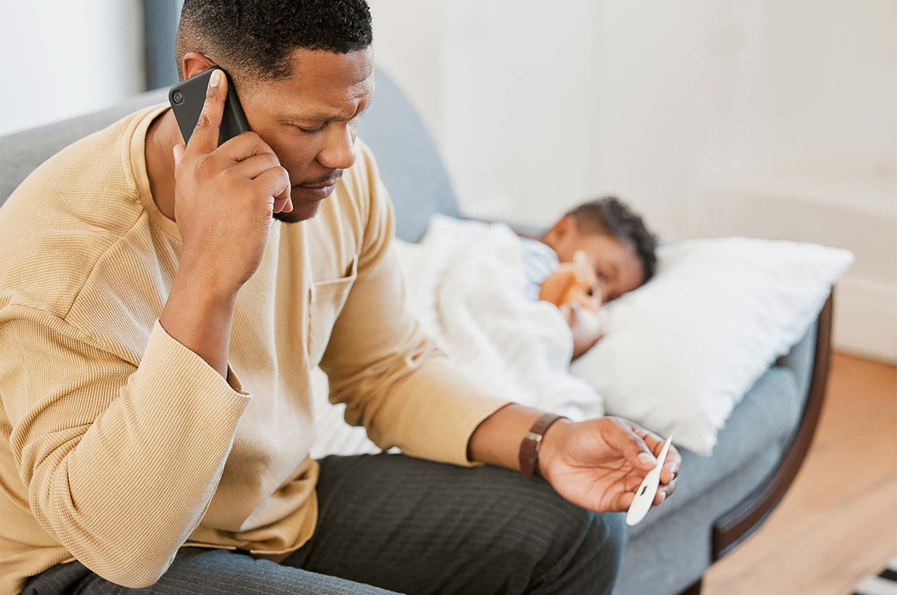 A father calls urgent care for fever, looking at a thermometer with his sick son on the couch.