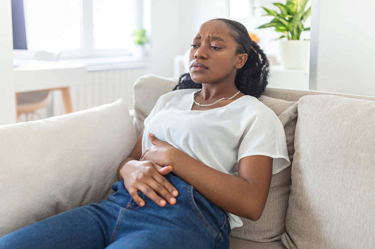 A young woman in a white shirt and blue jeans sits on a beige couch in a bright living room and holds her stomach, closing her eyes in discomfort from uterine fibroid symptoms.