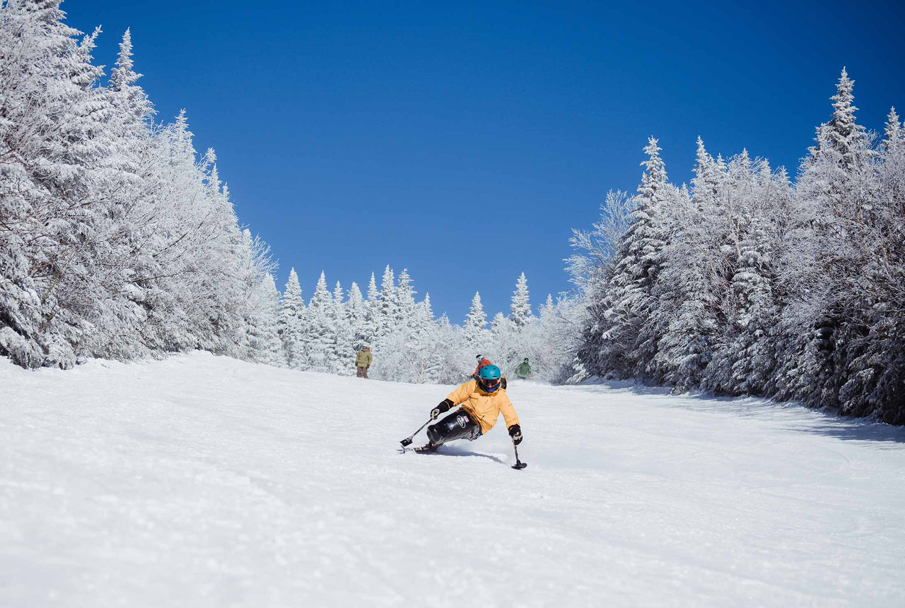 Chris Young, a five-time Paralympian and Spaulding Adaptive Sports Centers coach wearing a bright yellow jacket and blue helmet, is in a Mono-ski on the slopes.