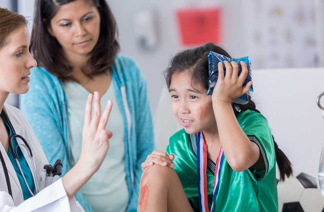 A little girl in a team jersey, with a medal around her neck, holds a cold pack to her head. Her cheek and shin are smeared with blood. A doctor holds up three fingers to test her vision while her mother looks on.