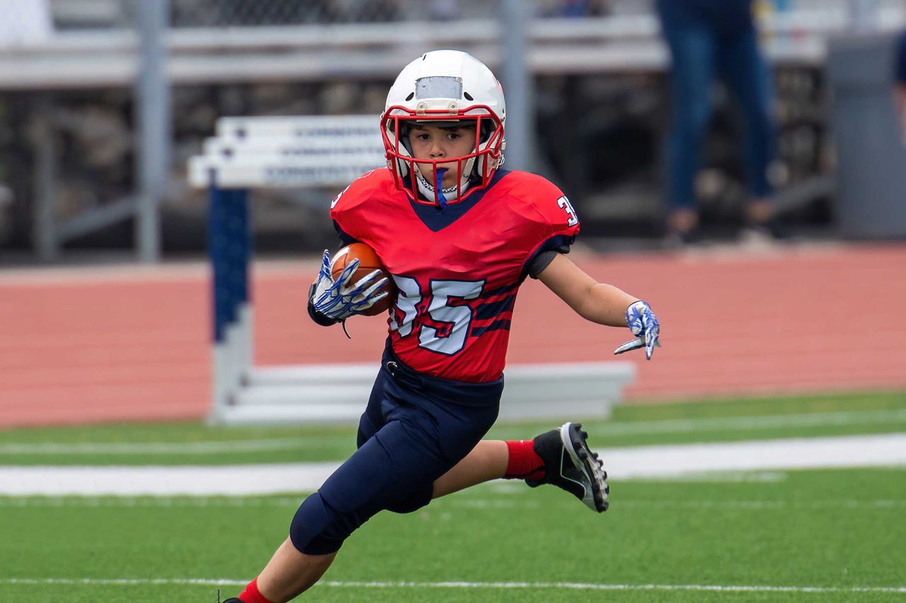A young boy in football gear runs on a football field with a football.