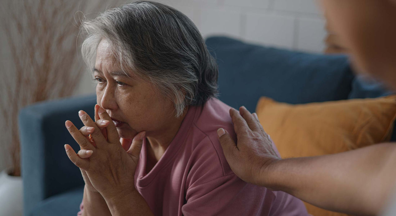 A person' reaches out their right arm to comfort an older woman in a pink t-shirt on her shoulder as she sits looking worried with both hands clasped by her mouth