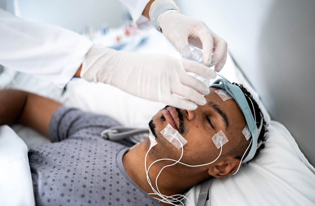 A doctor wearing white gloves adjusts tubes on a man participating in a sleep study