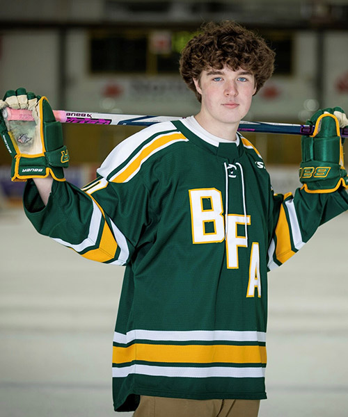 A high schooler wearing their green hockey gear and holding a hockey stick behind their upper back