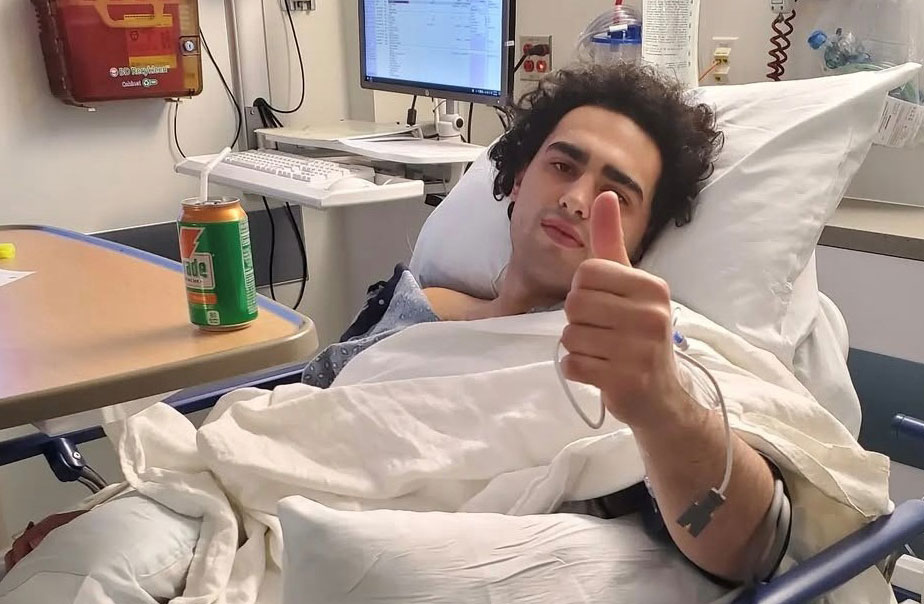 Angelo, a teenaged boy, gives a thumbs up as he lies in his hospital bed.