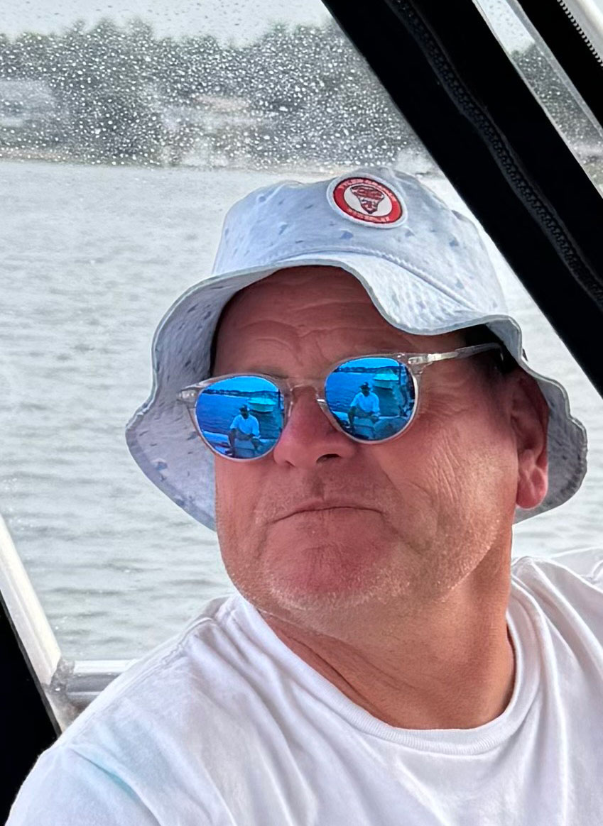 Bob Foley, a 61-year-old white man in a bucket hat, white T-shirt, and mirrored sunglasses, sitting by the window on a boat.