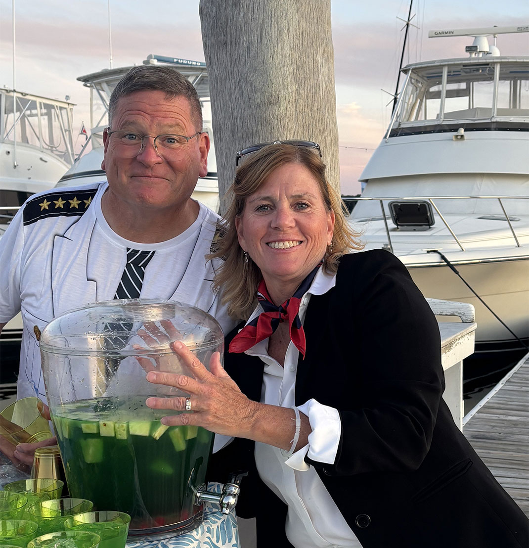 A middle-aged white couple poses by a table of drinks on a dock with a boat in the background.