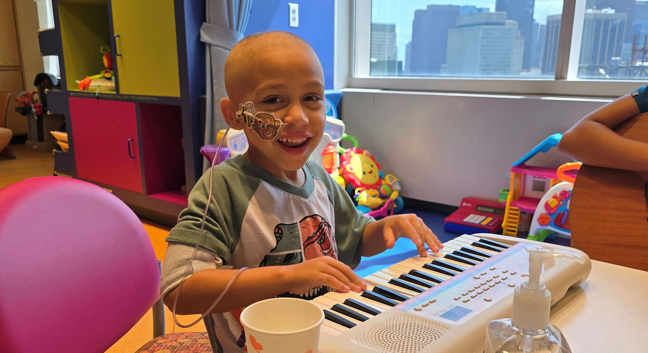 In a brightly colored hospital playroom full of toys, a small boy plays on a keyboard. He's bald, and a tube goes into his nose, held in place on his cheek by cartoon stickers.