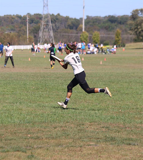 A young woman wearing a white jersey with the number '16' is actively playing ultimate frisbee on a grassy field. The setting is an open outdoor area with scattered players and spectators in the background. The scene captures dynamic movement and a casual sports atmosphere under clear skies.