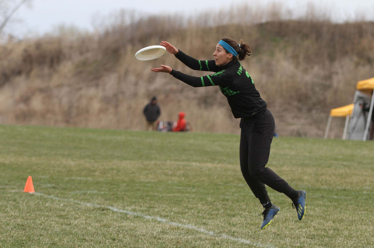 A person is captured mid-air while catching a frisbee on a grassy field. The setting appears to be an outdoor sports event with cones marking the play area. The individual is dressed in athletic attire, and the background includes a hill and a yellow tent. The scene conveys action and focus.