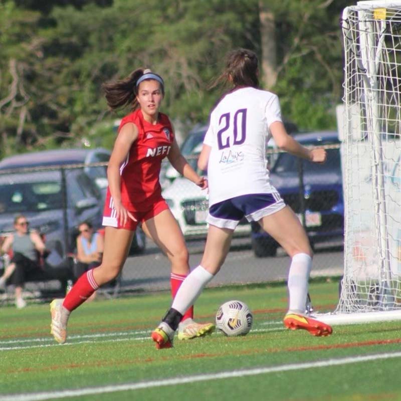 One high school girls soccer player, with a ponytail and headband dressed in a red team jersey and socks with grey and orange soccer cleats, plays against an opposing high school girls soccer player who is dressed in a white team jersey shirt, navy blue shorts with white trim, white socks, and orange cleats during a match; the soccer ball is on the ground between both players on the pitch