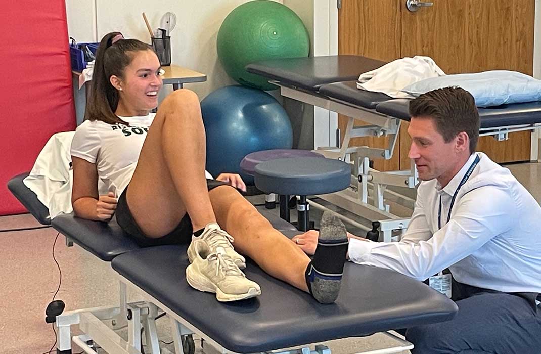 An athlete sits up halfway on a medical examination table during physical therapy as a doctor, who is seated, checks her left knee