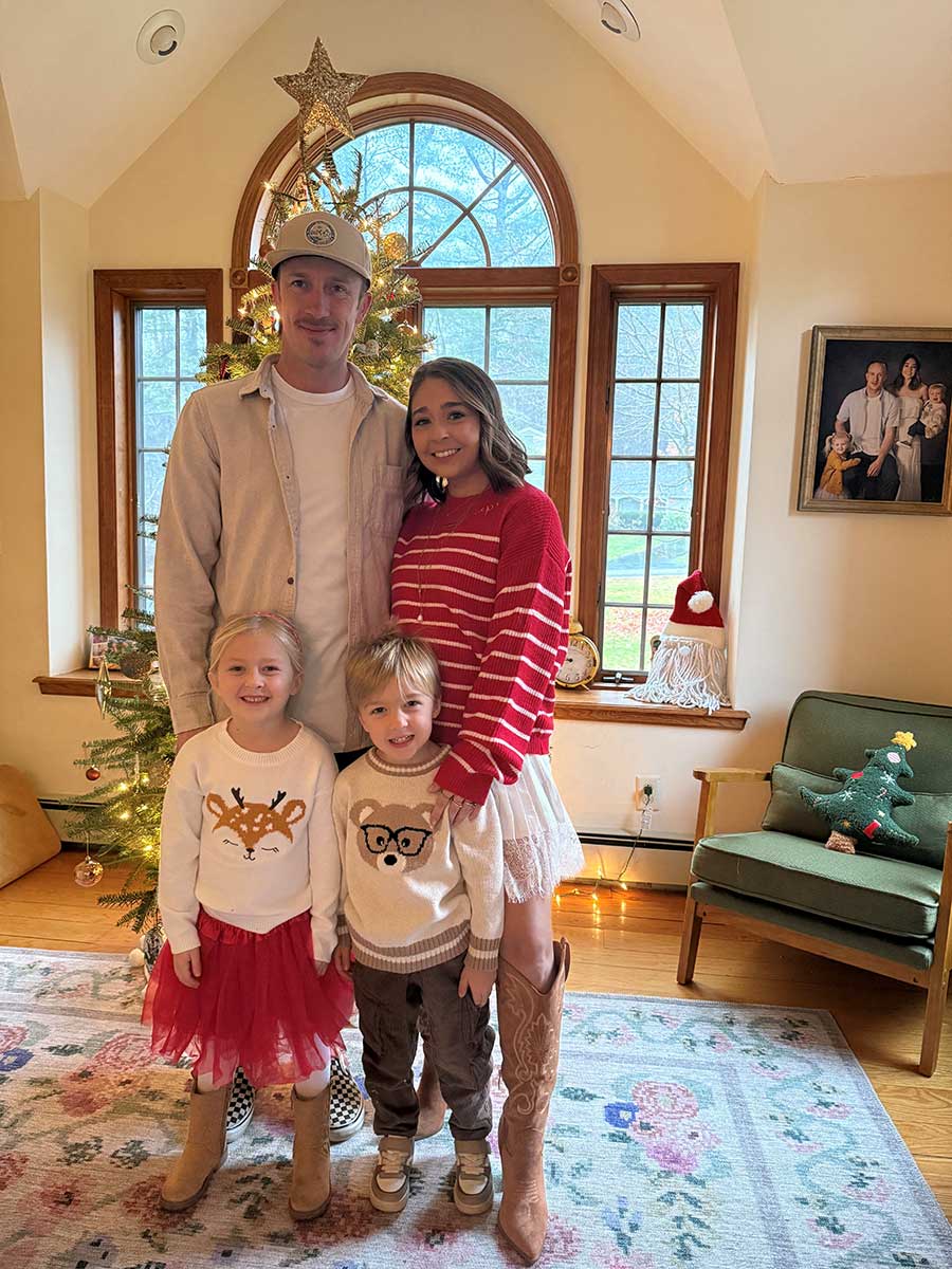 Kate, a white woman in her 30's wearing a red sweater, a frilly short skirt, and cowboy boots, poses in front of the family Christmas tree with her husband and two young children.