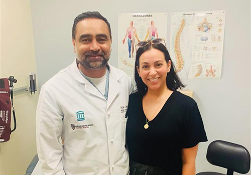 Patient and doctor smiling together in a clinic room. The patient wears everyday clothes, while the doctor stands beside her in a white coat. Medical equipment and exam table visible in the background.