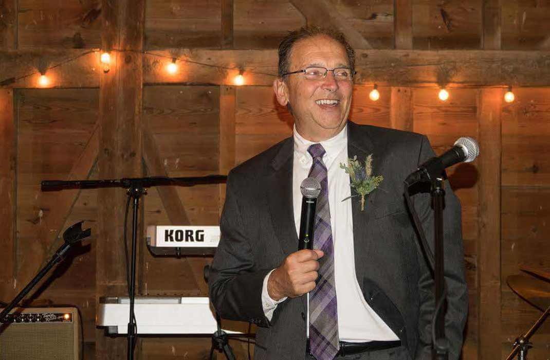 A man in a formal gray suit with a purple tie holds a microphone while standing in a rustic wooden venue. The setting features string lights and musical equipment, including a visible Korg keyboard.