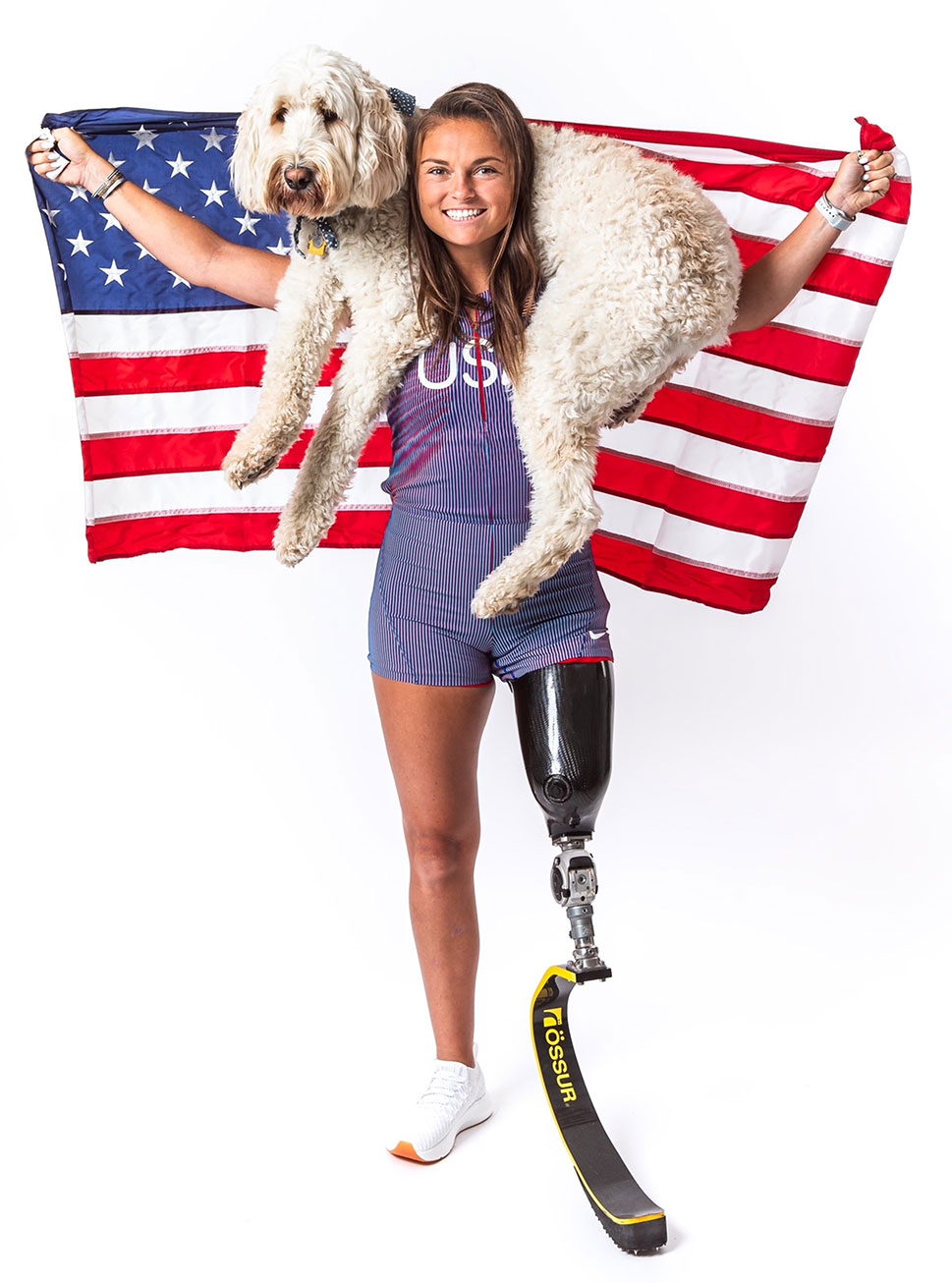 Noelle stands with her dog, a poodle mix, slung around her neck. She holds up an American flag and poses with her prosthetic leg in the foreground.