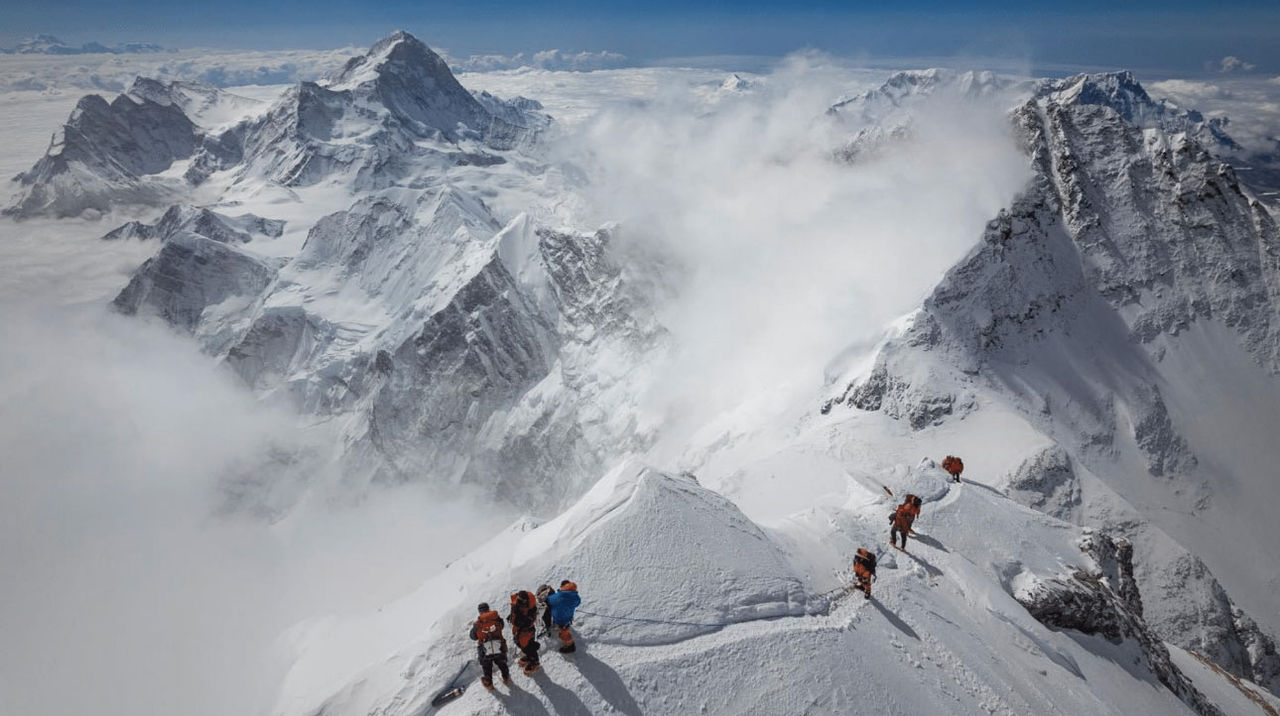 A dramatic shot of a ridge of Mt. Everest, seen from above. Mountaineers are making their way along a trail, dwarfed by the landscape.
