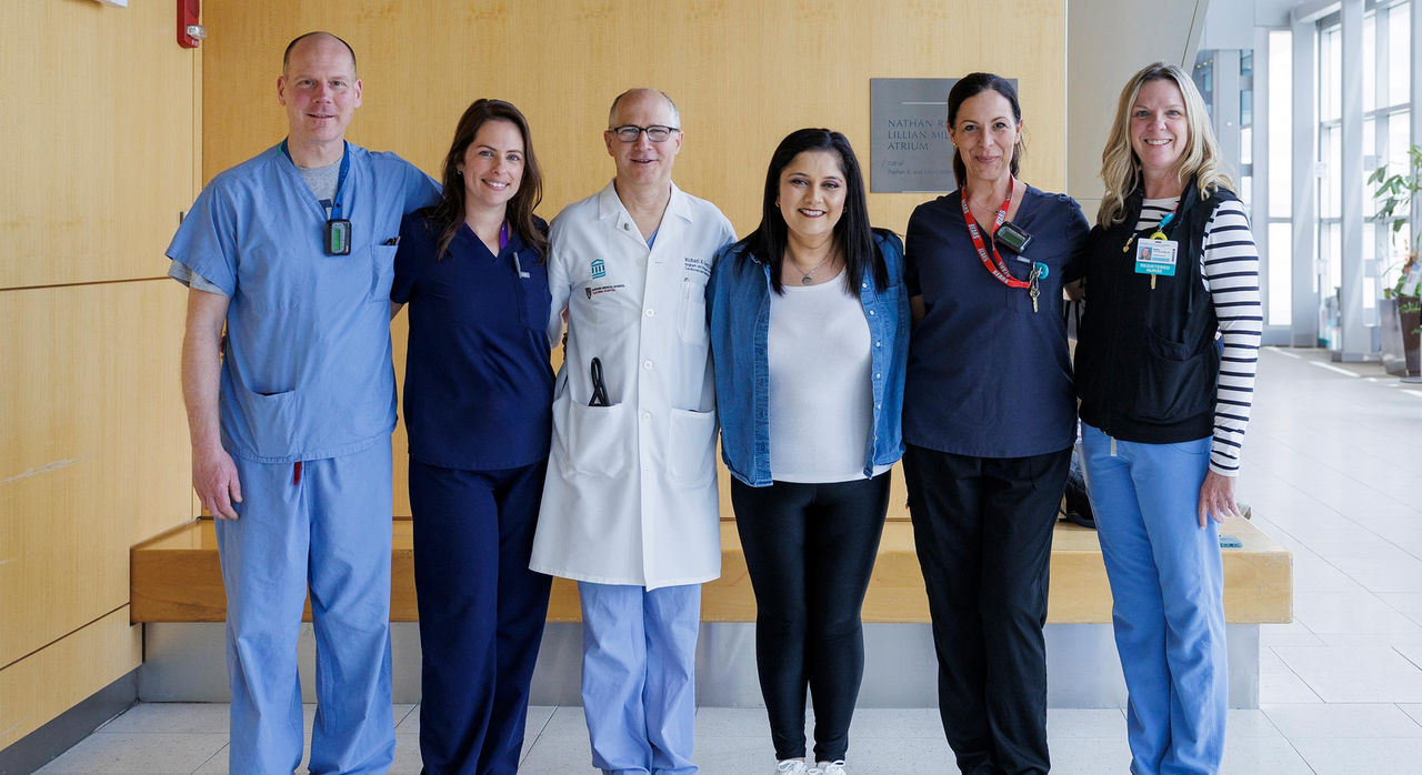 5 members of a patient care team in scrubs smiling and standing with a patient
