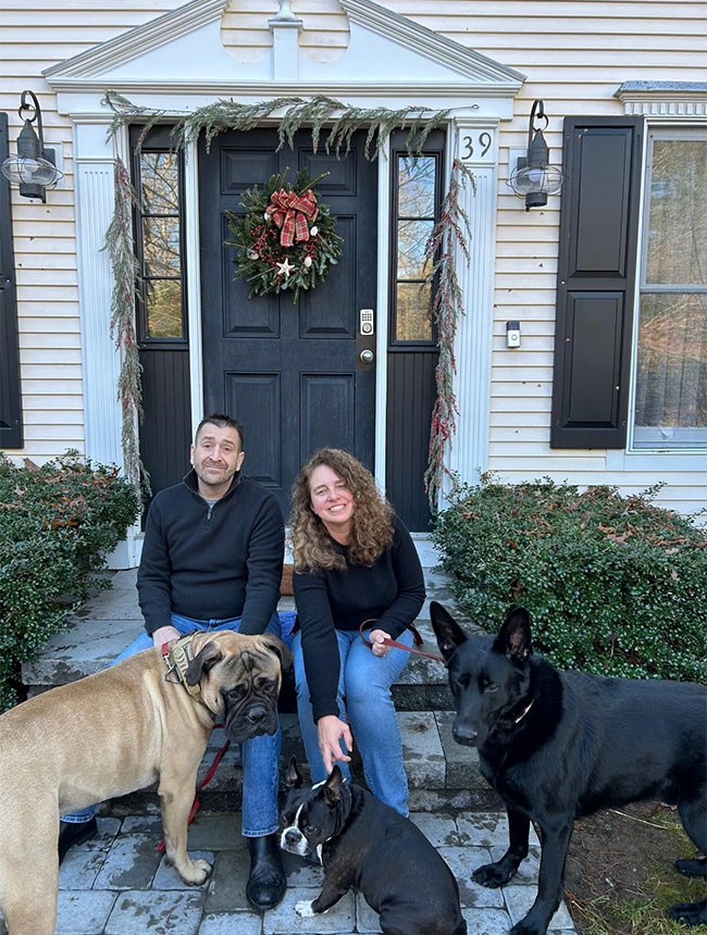 A man and woman sit on house steps during the day with three dogs of varying breeds and sizes such as a Bull Mastiff, a Boston Terrier, and a black German Shepherd. The door behind them features holiday decorations.