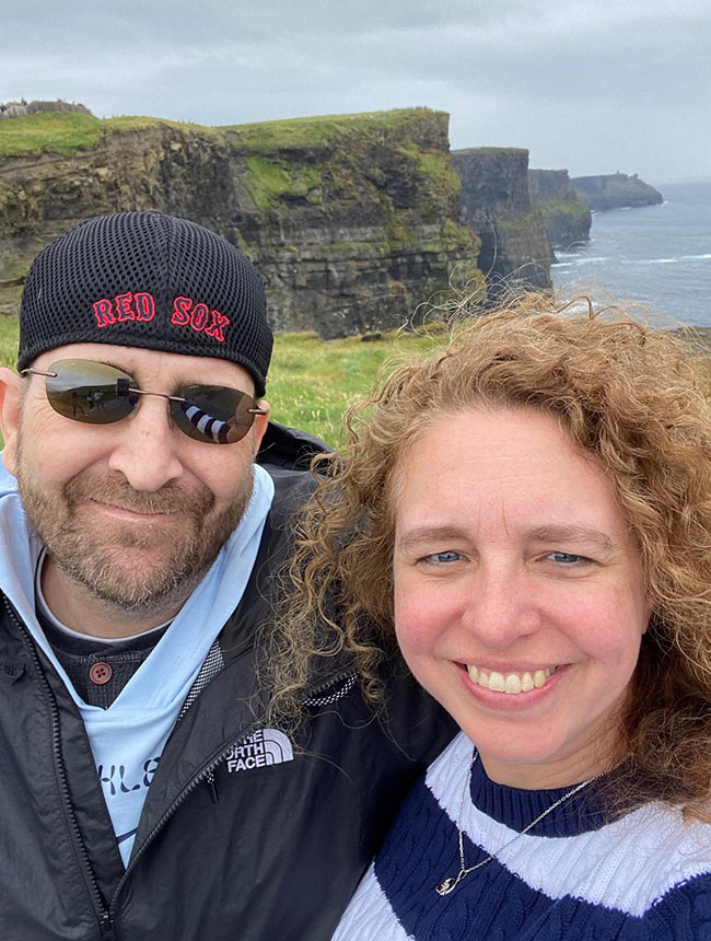 Two adults dressed in casual, layered clothing suitable for cool weather are pictured in the foreground, standing outdoors with dramatic sea cliffs in Ireland. The outdoor setting is overcast, with lush green grass atop the cliffs and rugged rock formations visible.