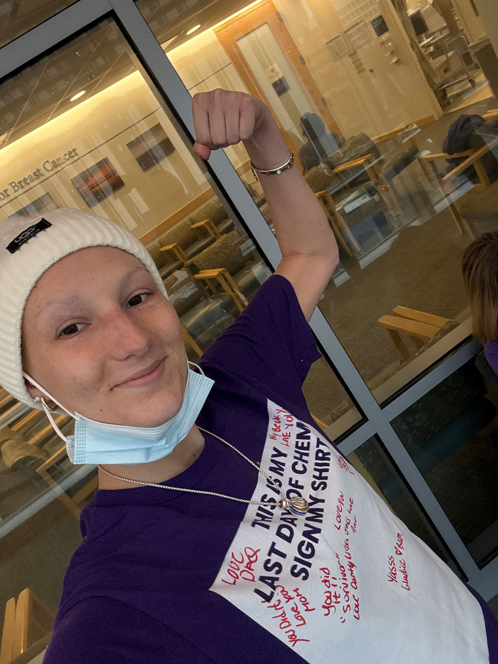 A woman is flexing her arm while wearing a dark blue t-shirt that celebrates her last day of chemotherapy. The shirt features signatures and encouraging messages from friends and family. She is also wearing a beanie and a mask. In the background, there is a waiting area with chairs and a sign for a cancer center.