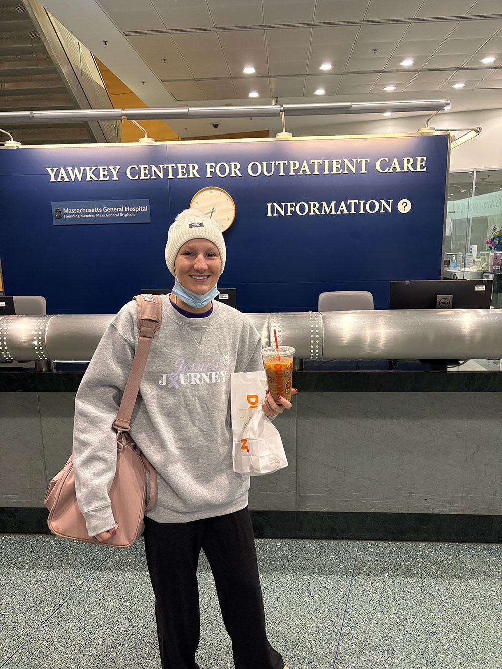 A woman stands in front of the Yawkey Center for Outpatient Care at Massachusetts General Hospital, holding a Dunkin iced coffee in one hand and a takeout bag in the other. She is wearing a gray sweatshirt with "Jessie’s Journey" printed on it and casual black pants. A pink bag is slung over her shoulder. The background features a reception area with a sign indicating information services.