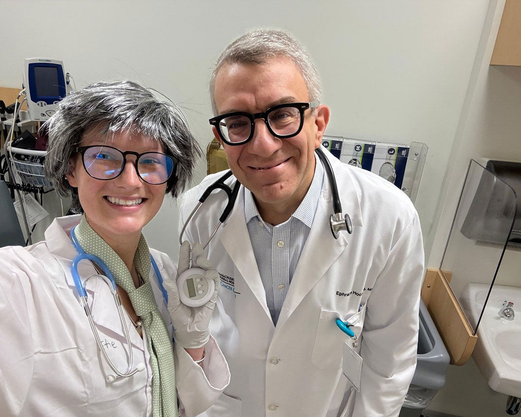A woman on the left is dressed in a costume imitating a doctor, complete with a gray wig, lab coat, stethoscope, and a scarf. She is playfully posing with a man on the right, who is also wearing a lab coat and stethoscope. The setting is a medical office, with medical equipment visible in the background.