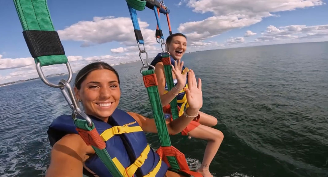 Two women are shown parasailing over the ocean, wearing life vests in bright colors. Both are raising their hand in a wave and smiling as they enjoy the experience. The background features blue skies with fluffy clouds and the calm water below.