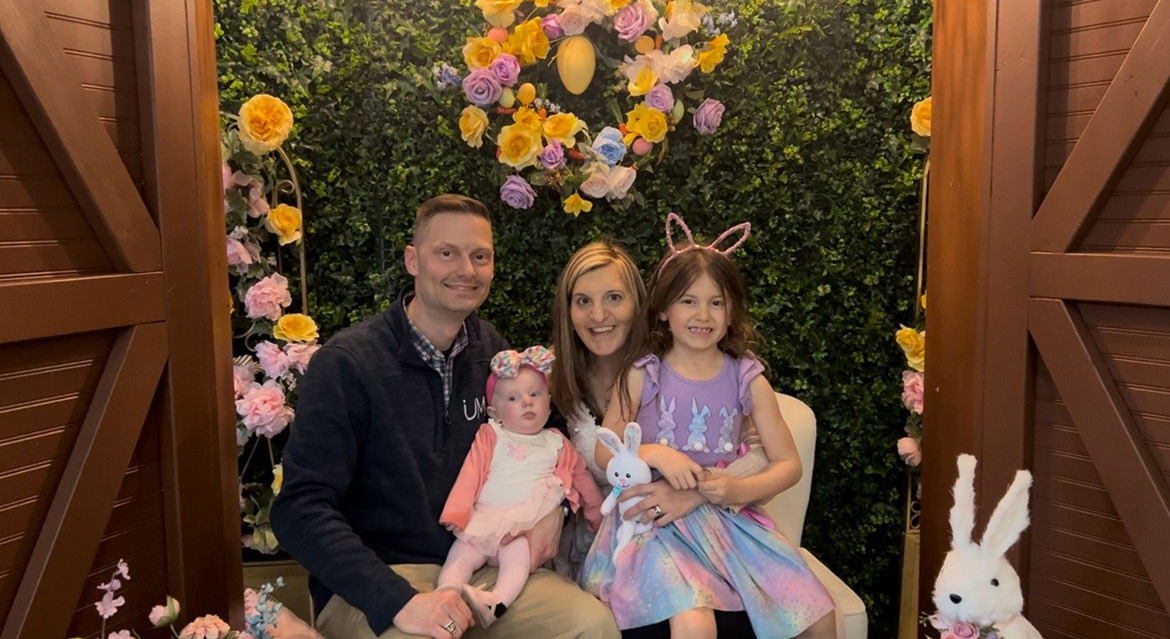 Stefania, her husband, and her two little daughters posing in a display of Easter flowers and bunnies.