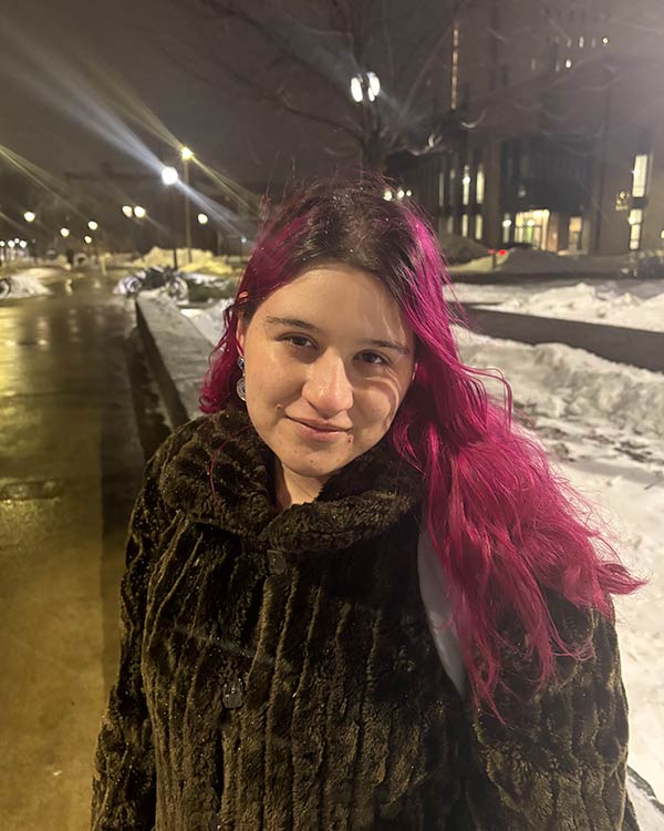 A person with vibrant pink hair and a dark coat is seen standing outside on a snowy night.