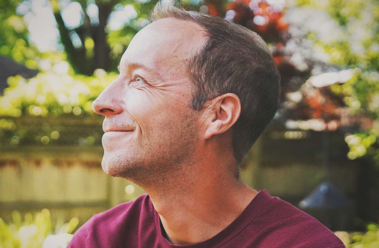 Side profile of smiling adult man outdoors on a sunny day.