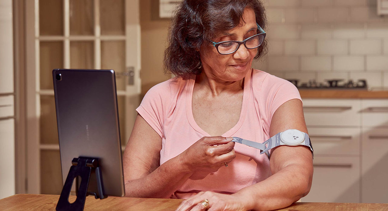 An older woman patient with brown hair, glasses, and a light pink top adjusts her health armband with a tablet in front of her