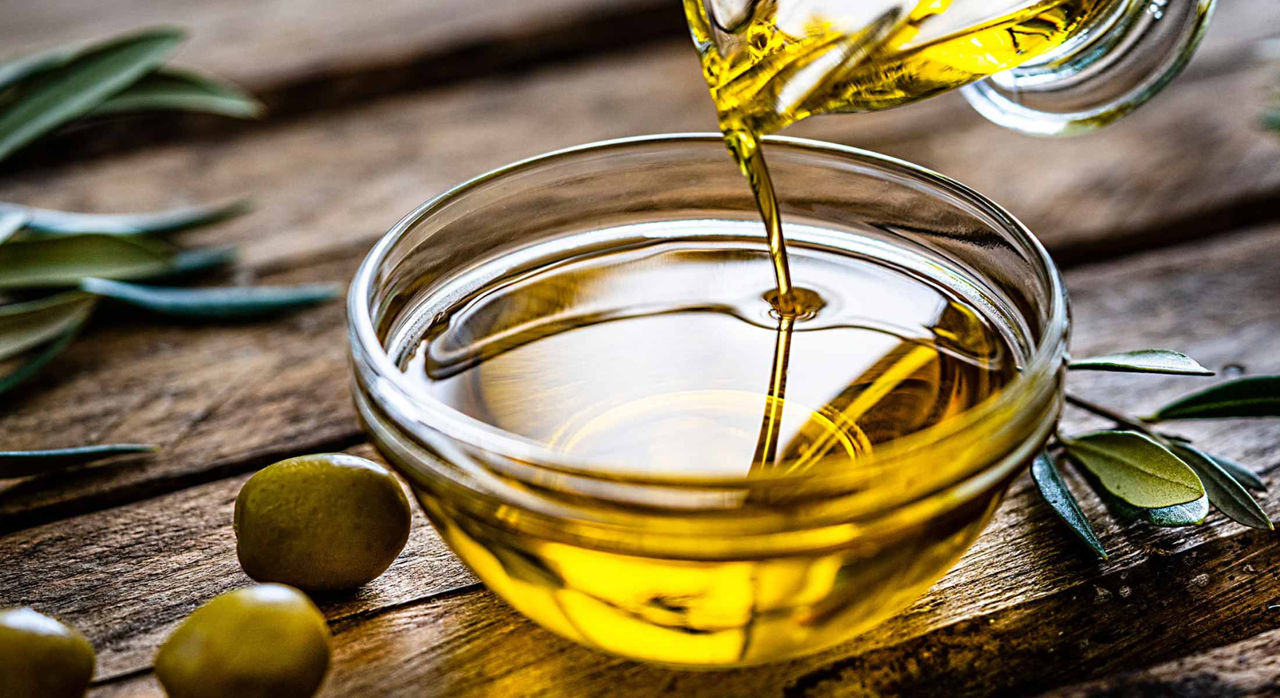 Olive oil poured into a glass bowl with olives and olive leaves on the table