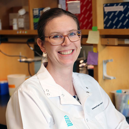 A scientist, wearing glasses and a white lab coat with the Mass General Brigham logo, smiles in the lab with shelves and materials in the background.