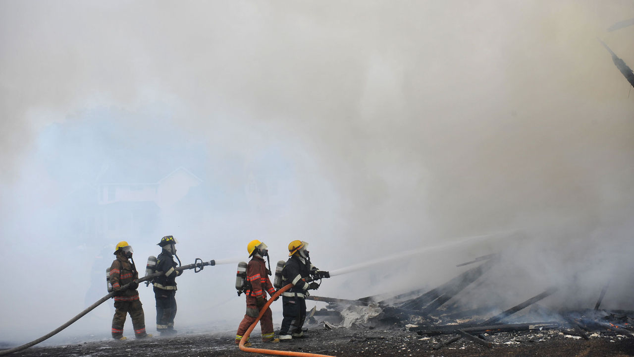 Four firefighters in firefighting gear aim hoses at the remains of a smoking building.