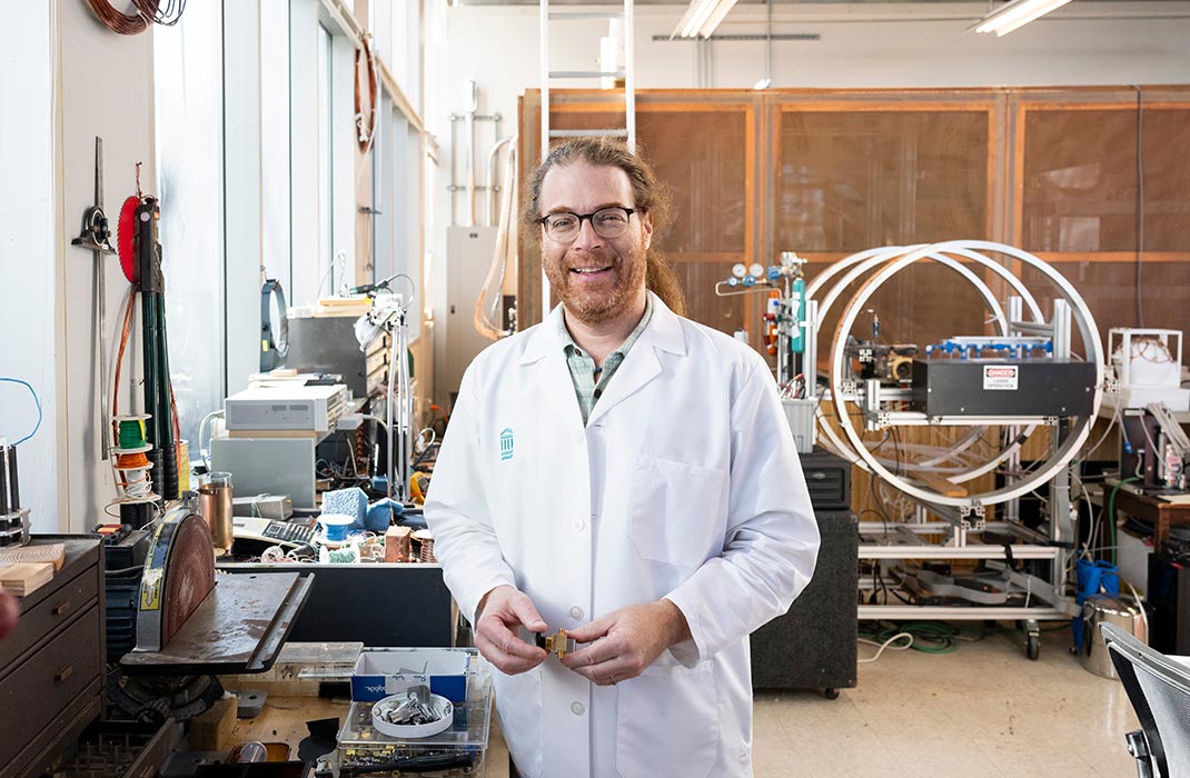 A person wearing a white lab coat stands in a well-lit laboratory filled with scientific instruments and technical equipment.