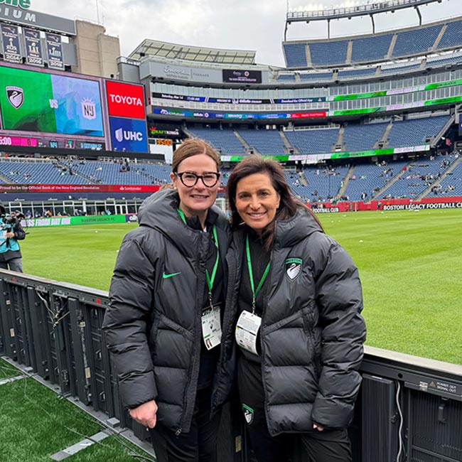 Two sports medicine doctors wearing black jackets with the Boston Legacy FC emblem while standing on the sideline of a soccer stadium.