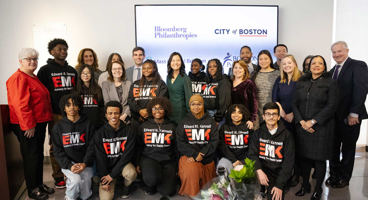 A group of Edward M. Kennedy Academy students in black t-shirts, with Mayor Michelle Wu, Dr. Anne Klibanski, and other leaders and representatives. There is a monitor behind the group with a slide featuring the logos of Bloomberg Philanthropies and the City of Boston.