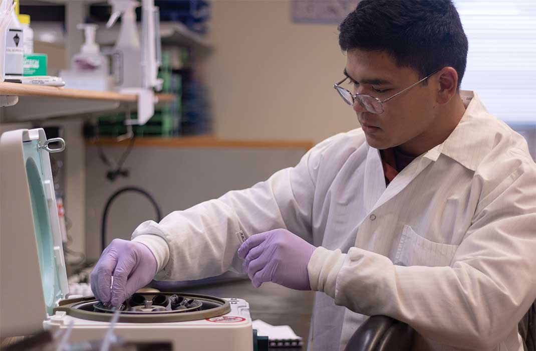 A research student in gloves and a white lab coat inserts vials into a centrifuge.