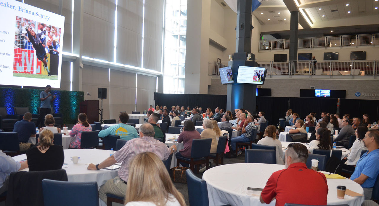 Attendees sitting at round tables looking at a projector screen during a conference