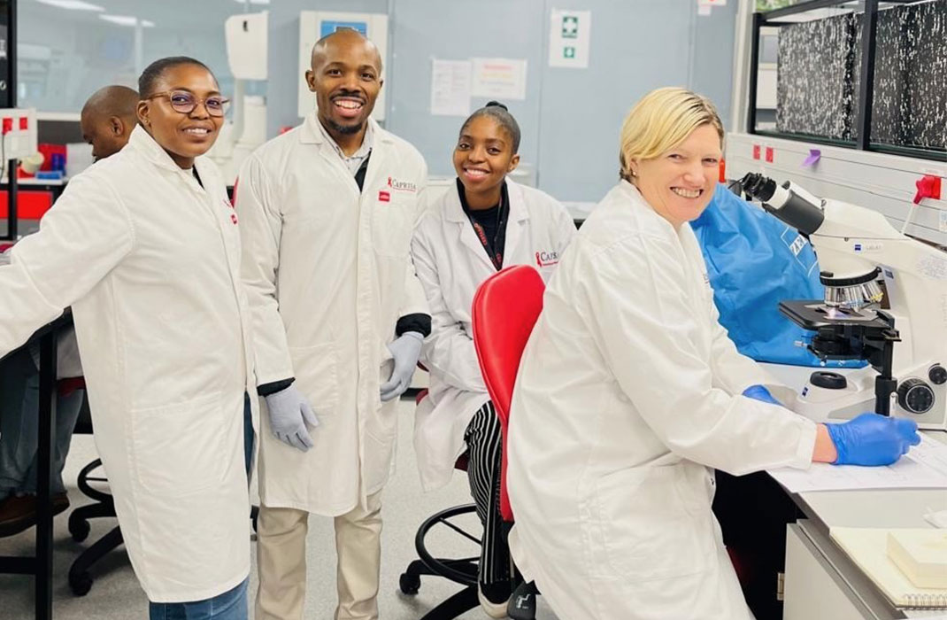 Four smiling researchers in white lab coats pose together in a lab. One investigator, Caroline Mitchell, sits in front of a microscope.