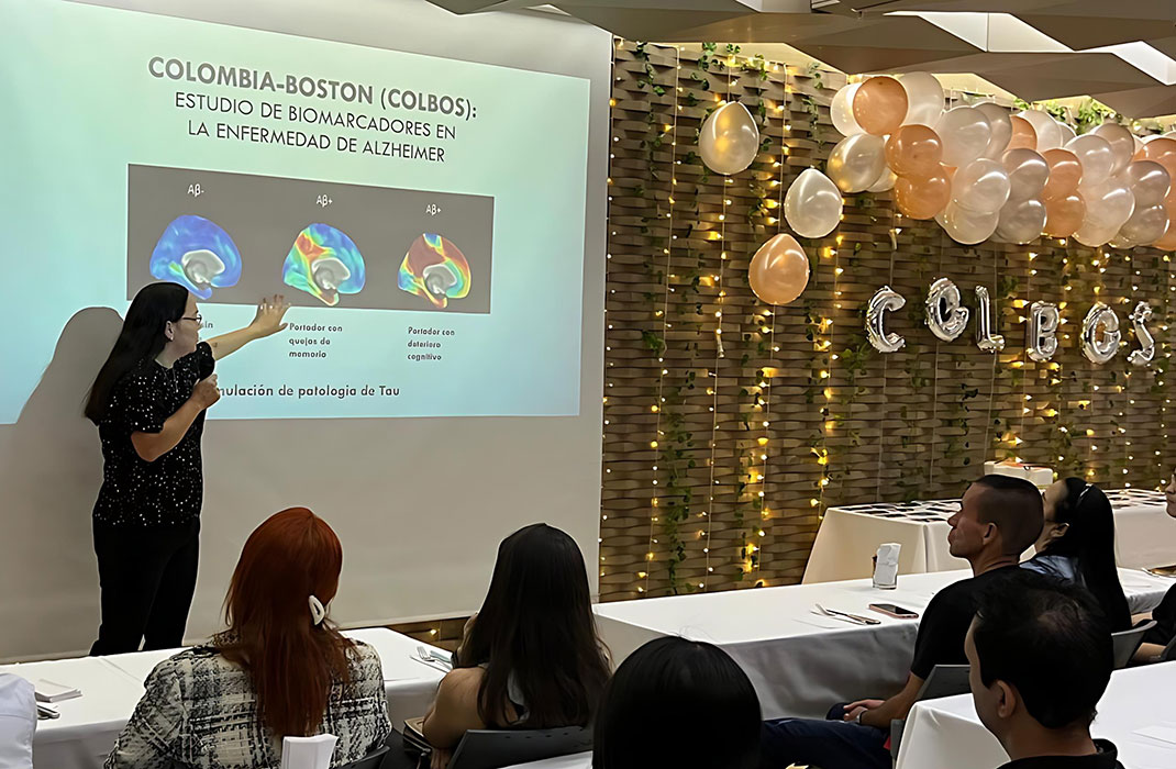 A woman gives a presentation to a roomful of people. To the right are champagne-colored party balloons and a line of silver balloons that read COLBOS.