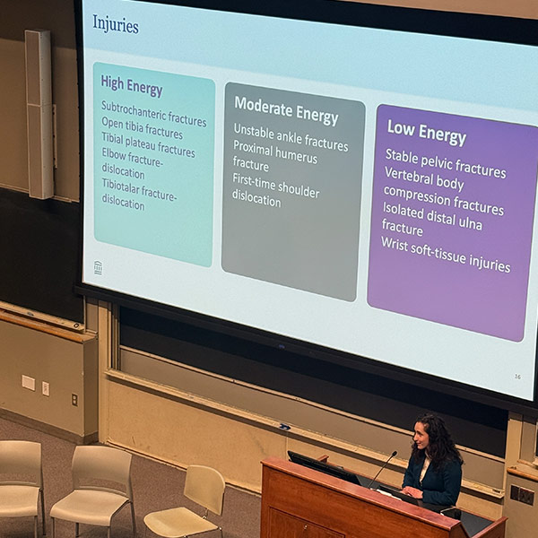 A presenter delivers a lecture on injury classifications in a university-style auditorium. The slide displayed categorizes injuries into high, moderate, and low energy, with examples listed under each category. The setting includes a podium, chairs, and a large projection screen.