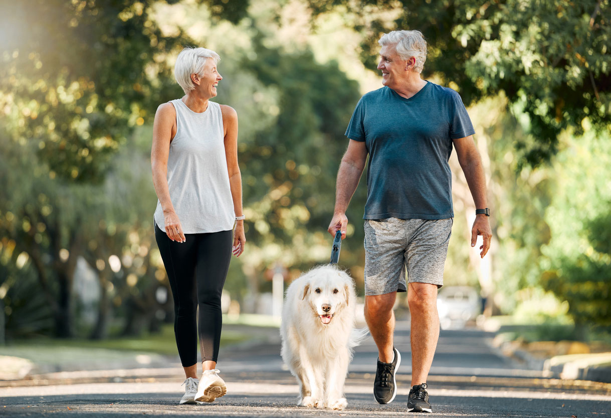 An older couple walking in the park with their dog.