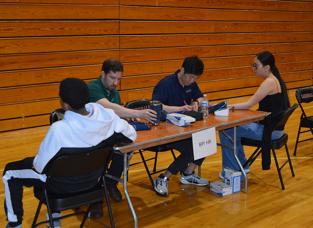 Two Mass General Brigham Sports Medicine team members sit at a table with two Boston Public Schools athletes. A sign on the front of the table reads, "BP/HR" for blood pressure/heart rate screenings.