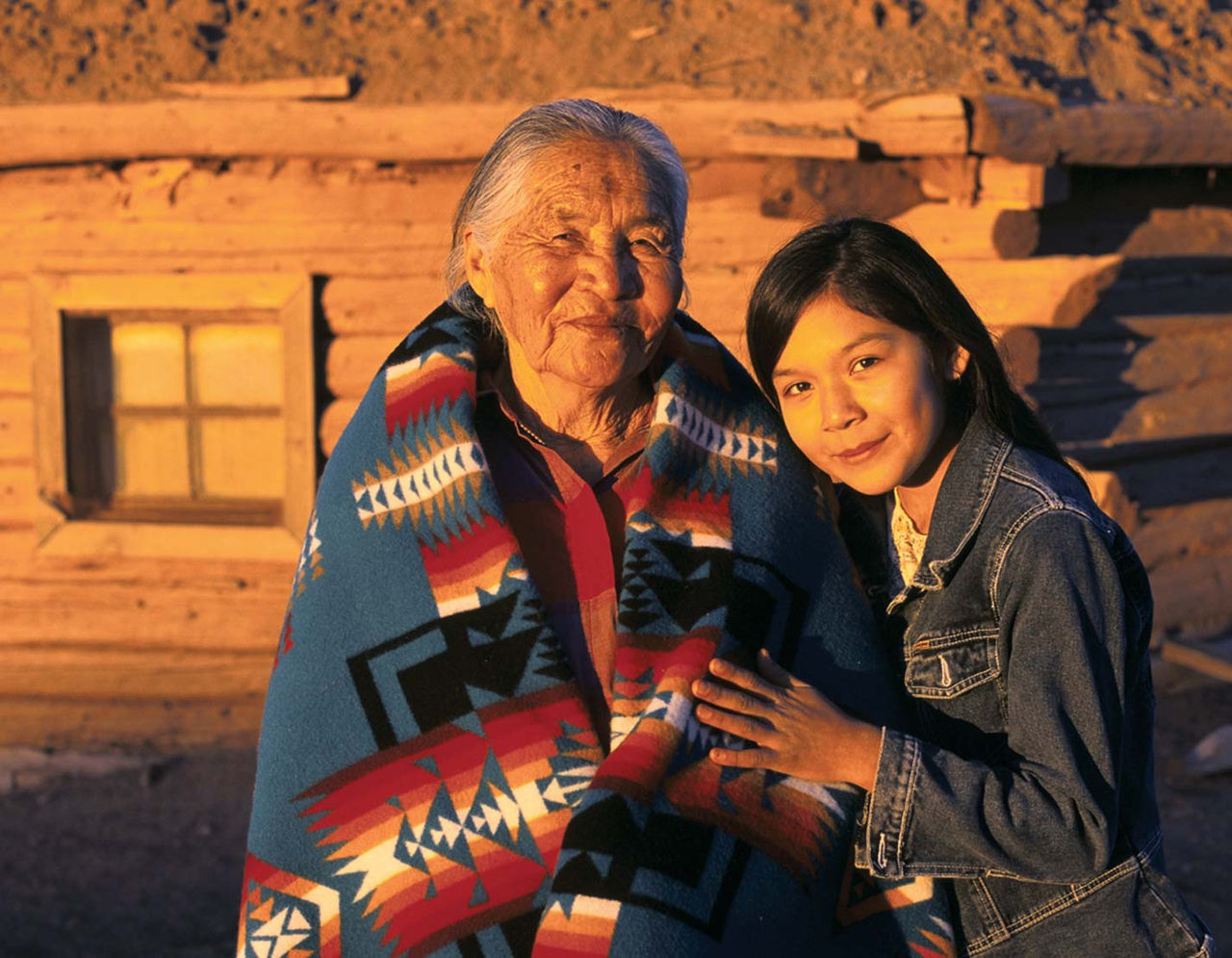 A Native American grandmother and granddaughter in front of a traditional house