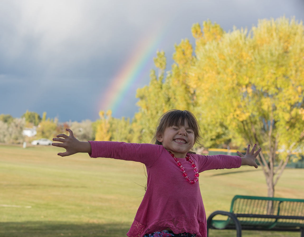 A child smiling with outstretched arms, in a pink long-sleeve top and a necklace, at a park with a rainbow and tree in the background