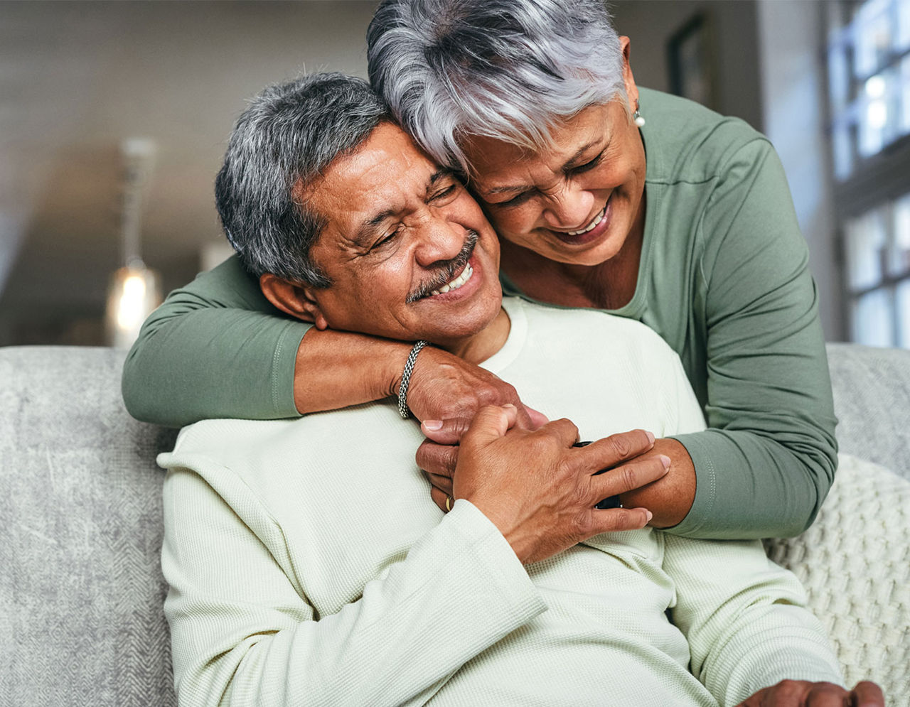 A man sits on a couch as a woman reaches over and hugs him.