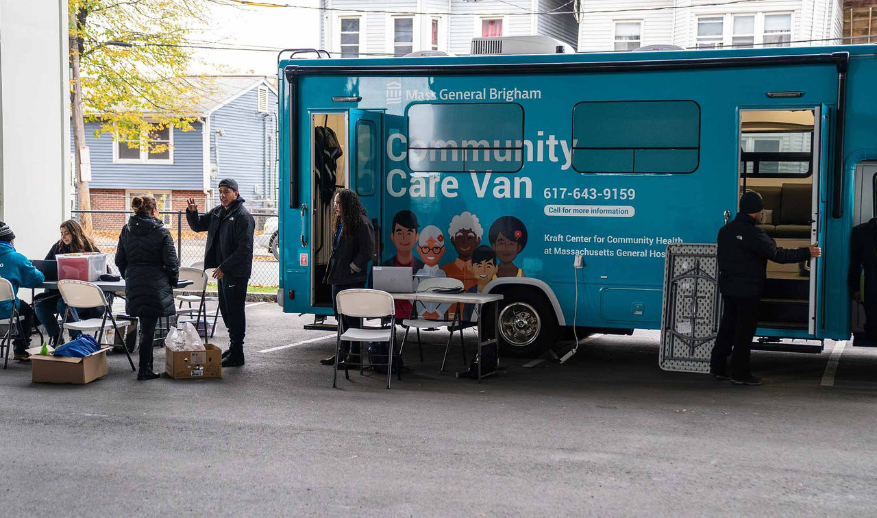A right side view of the Mass General Brigham Community Care Van parked at a location with tables, chairs, and boxes for set up. The van is wrapped in teal on the side and blue on the back consistent with the Mass General Brigham brand colors. The side panel has the Mass General Brigham logo on top with the words "Community Care Van" in large font and a phone number in smaller font. Part of the side wrap design includes 5 illustrated individuals of varying genders, ethnicities, and ages.